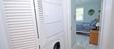 Una lavandería con lavadora y secadora de carga frontal, piso de madera y una puerta que da a un dormitorio con cama y mesita de noche.