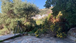 A stone-paved patio with a swing set, surrounded by lush greenery and yellow flowers. - Appartement Climatisé, RDC Villa, Avec Jardin Arboré. Proche Centre Ville Bastia (Ville-di-Pietrabugno)
