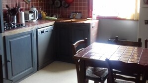A kitchen with a tiled backsplash, a sink, a stove, and a dining area with a table and chairs. - Appartement Climatisé, RDC Villa, Avec Jardin Arboré. Proche Centre Ville Bastia (Ville-di-Pietrabugno)