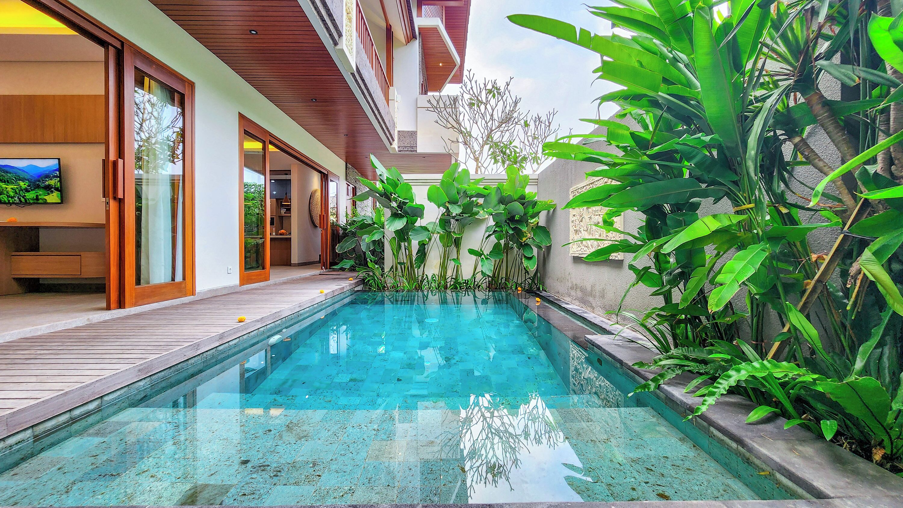 A pool area with a wooden deck, surrounded by lush greenery and a modern building with large windows and doors.