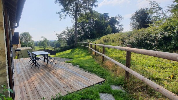 Eine Holzdar Terrasse mit Tisch und Stühlen, mit Blick auf eine grüne Fläche mit Bäumen und einem Zaun.