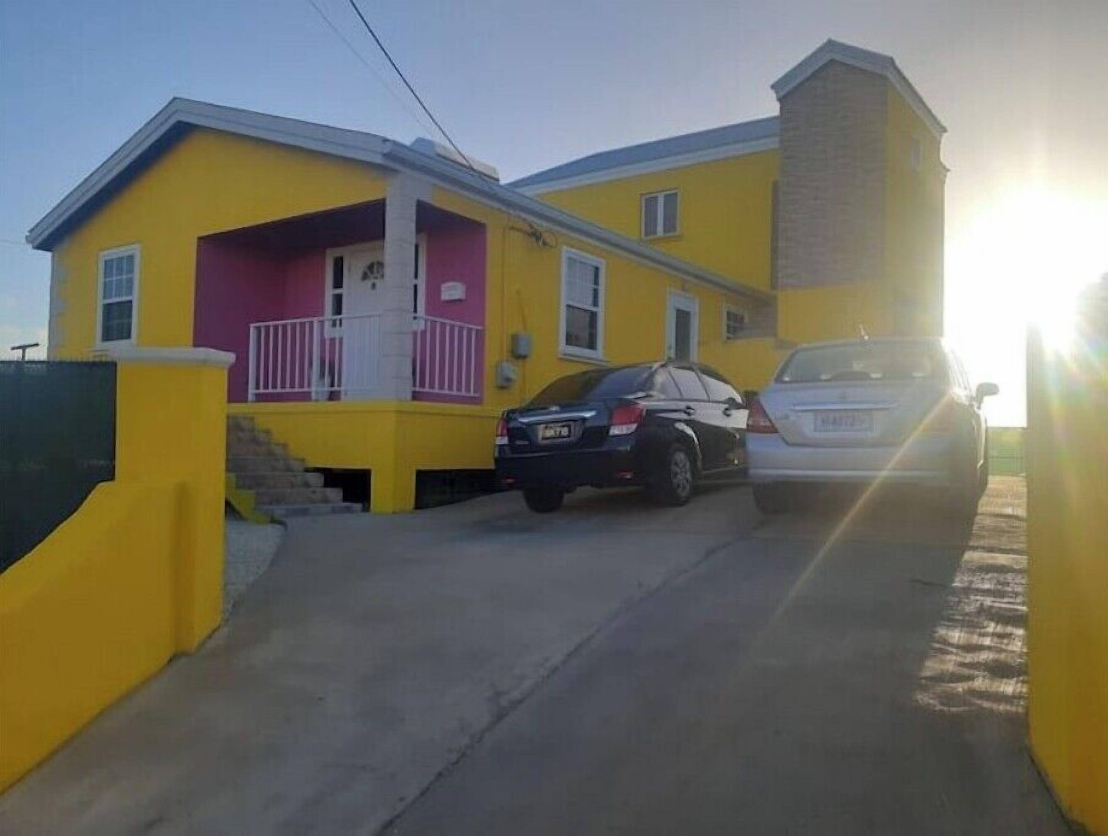 A yellow building with a pink front porch, two cars parked in front, and a clear sky.