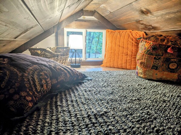 Ein gemütliches Schlafzimmer im Dachgeschoss mit schrägen Wänden, einem großen Fenster, einem gemusterten Teppich und einem farbenfrohen Kissen.