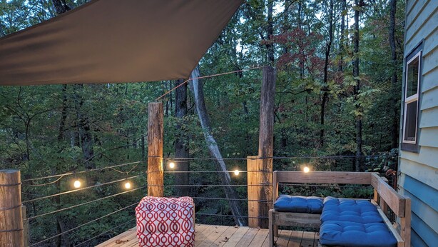 Eine Holzterrasse mit überdachter Sitzgelegenheit, Lichterketten und Blick auf einen bewaldeten Bereich.