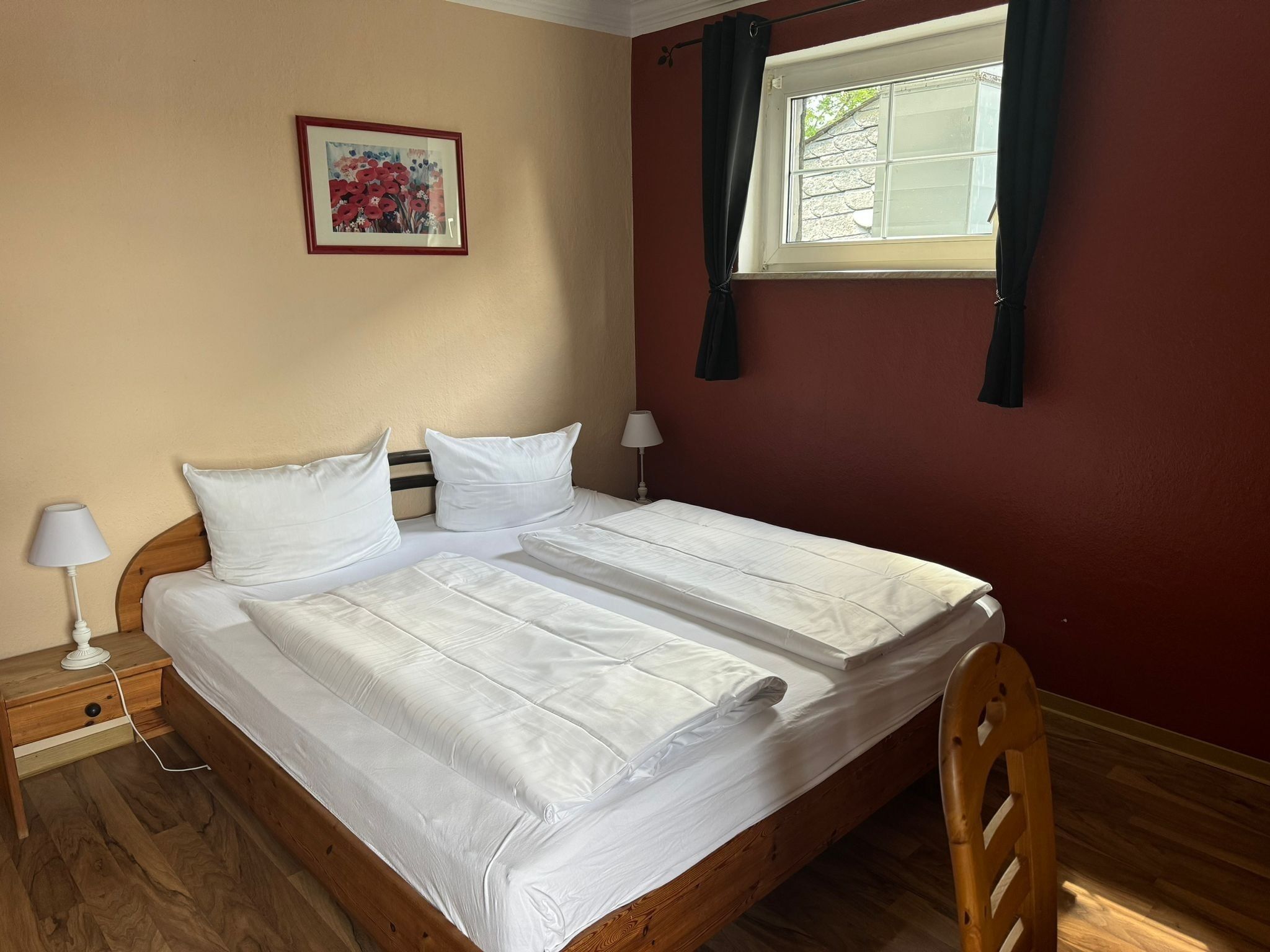 Un lit double avec du linge de lit blanc, une tête de lit en bois, une table de chevet avec une lampe et une fenêtre avec des rideaux.