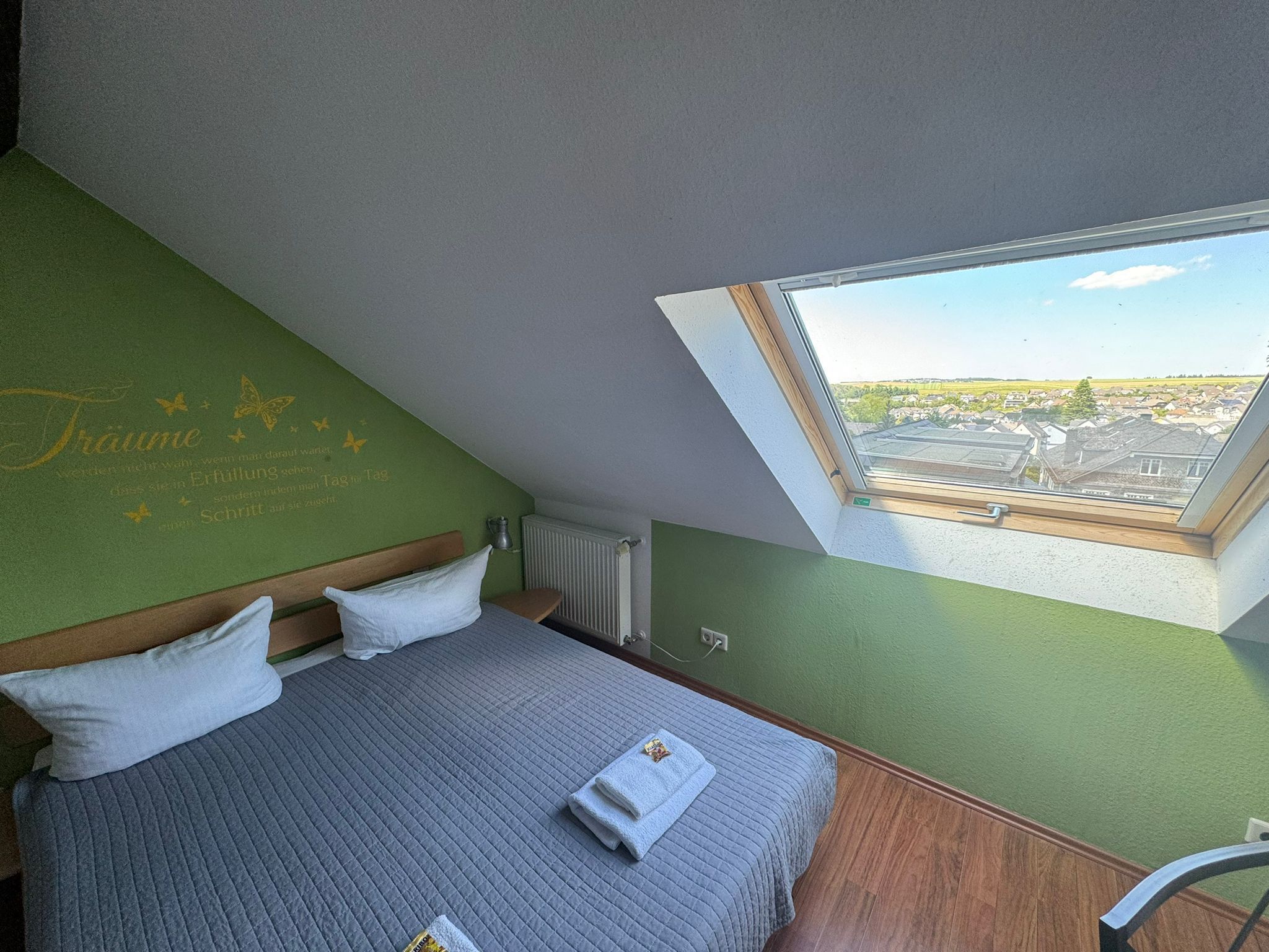 Ein Schlafzimmer mit schrägem Dach, einem Bett mit Kissen, einem Fenster mit Blick auf eine Wohngegend und einem Heizkörper.