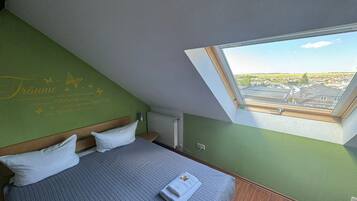 Ein Schlafzimmer mit schrägem Dach, einem Bett mit Kissen, einem Fenster mit Blick auf eine Wohngegend und einem Heizkörper.