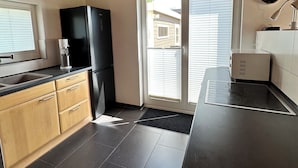 A modern kitchen with wooden beams, a black refrigerator, a stove, and a window with blinds. - Cozy vacation apartment, fully equipped 80 sqm with garden and underfloor heating (Attendorn)