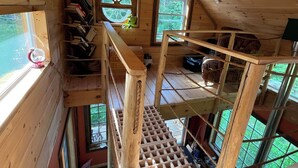 En træhytte med et soveværelse på loftet, en lille balkon og et vindue med udsigt over grøn vegetation.