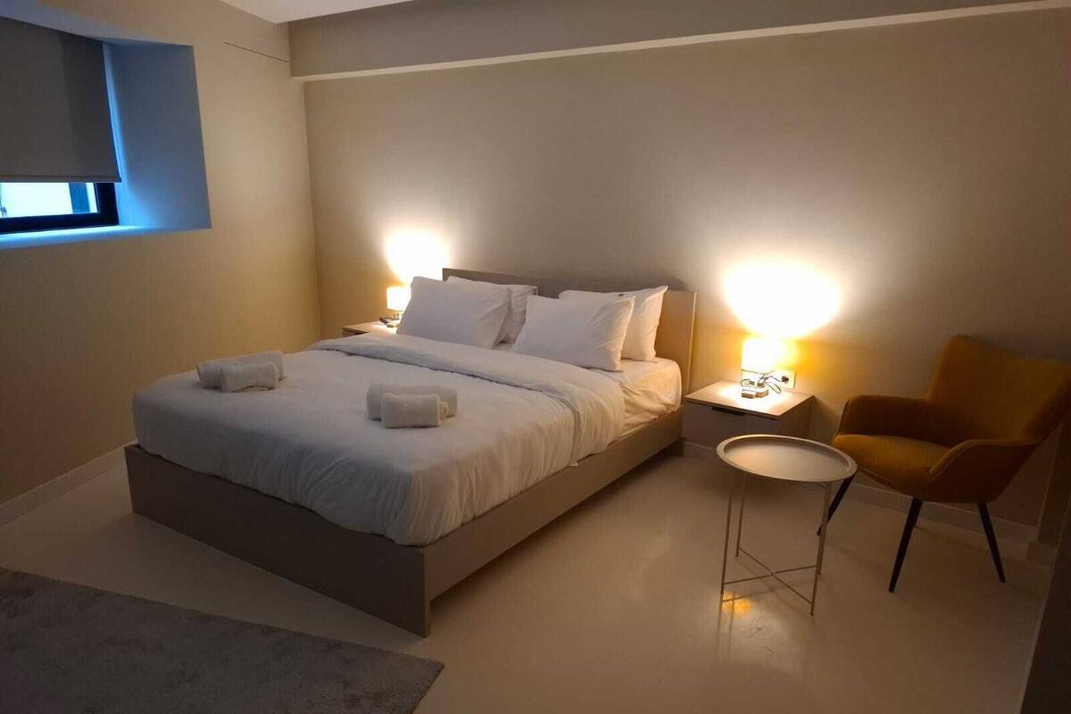 A modern bedroom with a large bed, bedside lamps, a small round table, and a chair.