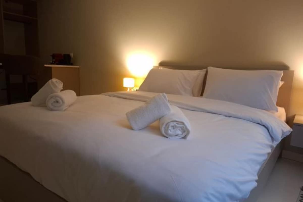 A neatly made bed with white linens and rolled towels, a bedside lamp, and a small table with a lamp on it.