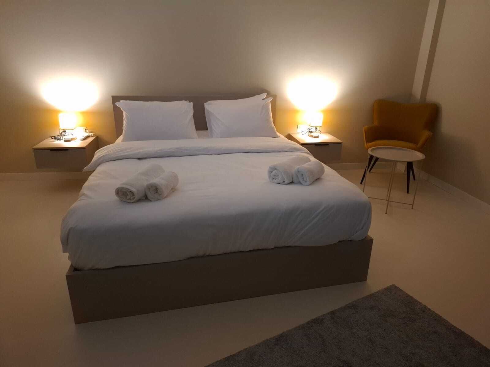 Ein ordentlich bezogenes Bett mit weißen Leinen und aufgerollten Handtüchern, zwei Nachttischlampen, ein kleiner runder Tisch und ein einzelner Sessel in einem modernen Hotelzimmer.
