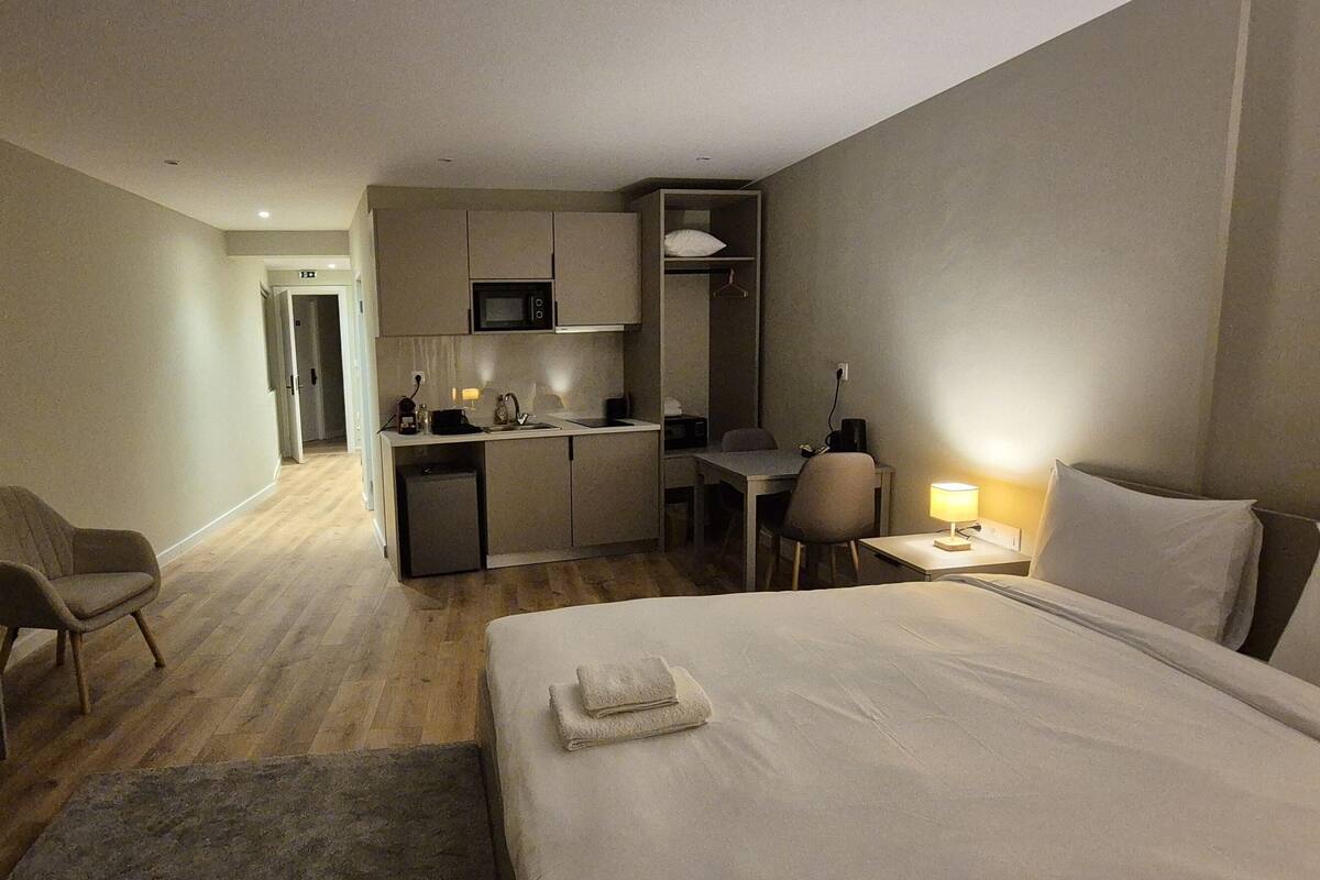 A hotel room with a bed, a desk, a chair, a television, a kitchenette, and a small table with a lamp.