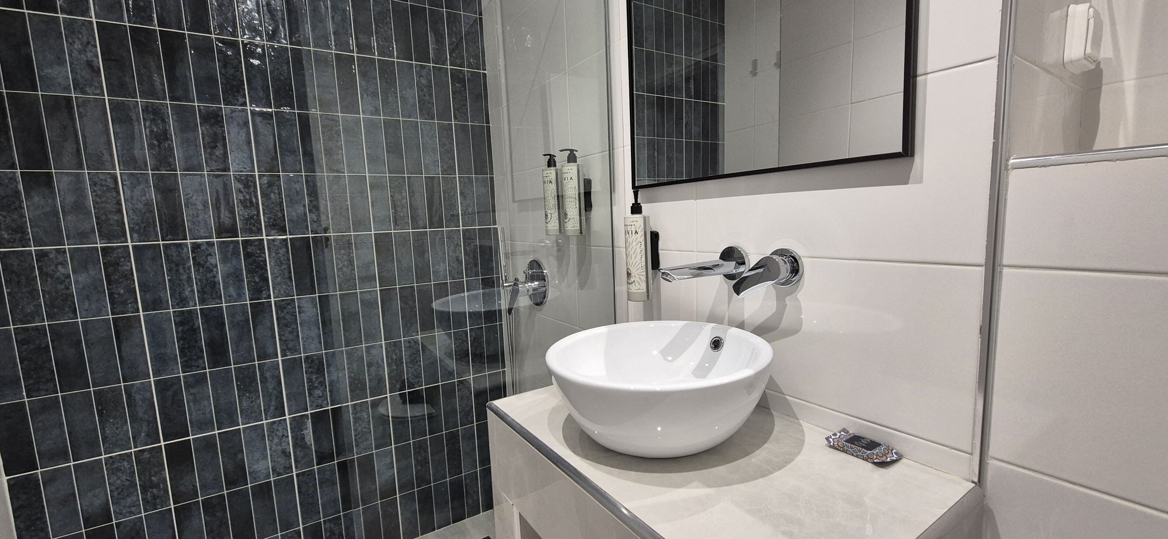 A modern bathroom with a large mirror, a white sink, and a shower area with dark tiles.