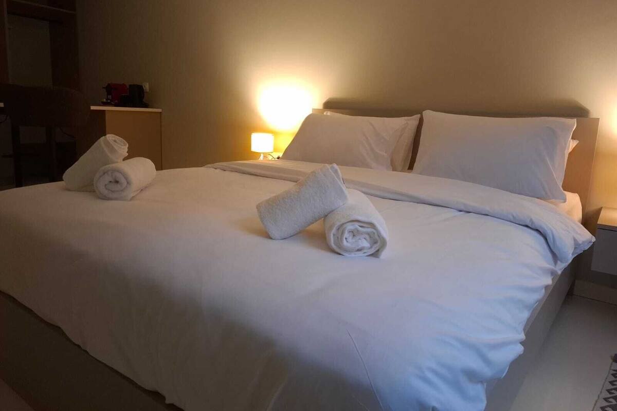 A neatly made bed with white linens and rolled towels, a bedside lamp, and a nightstand with a lamp.