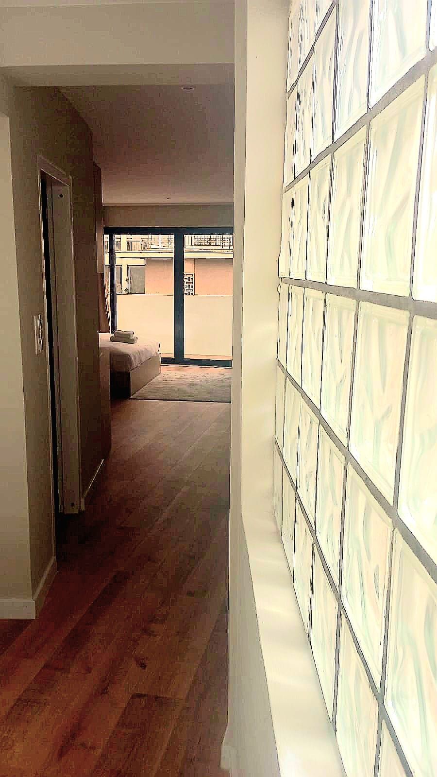A hallway with a wooden floor, a door on the left, and a wall with patterned glass panels on the right.
