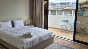 En pænt redt seng med hvide sengetøj og et foldet håndklæde, en senget lampe og udsigt til en balkon med en stol og et bord.