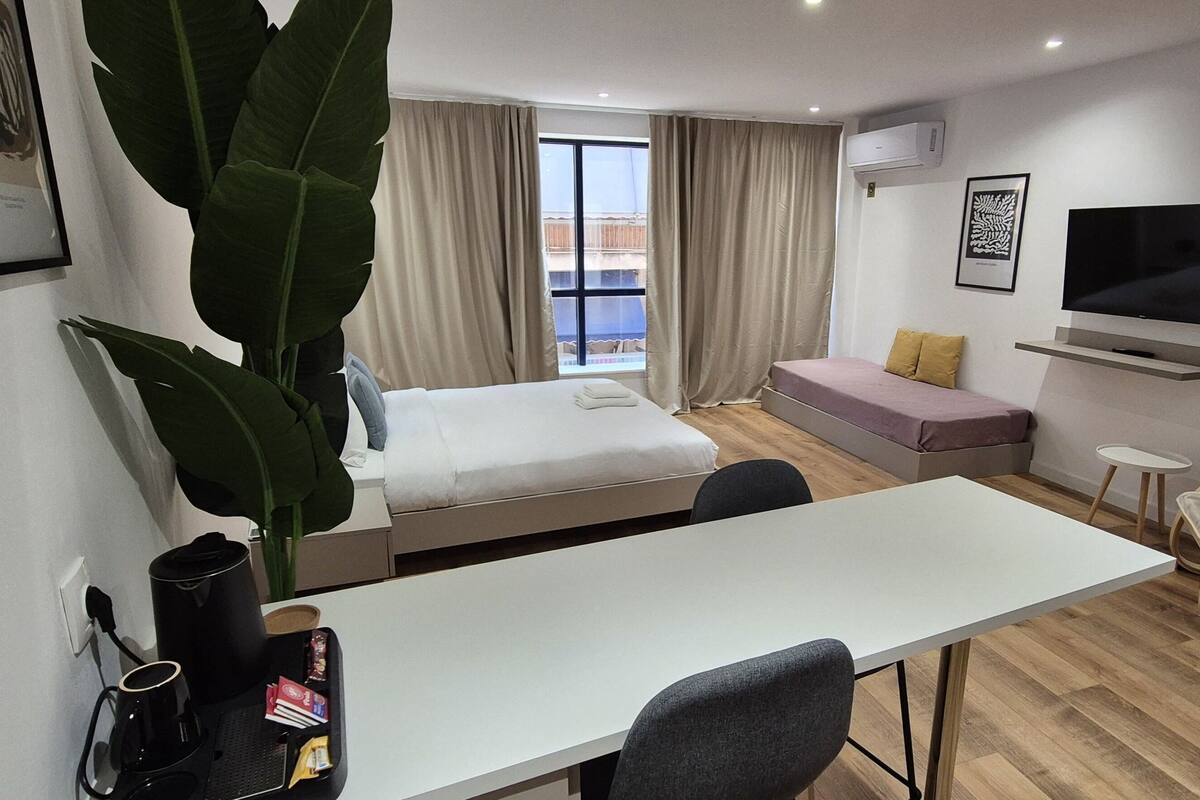 A modern hotel room with a bed, a desk, a chair, a TV, and a large plant.