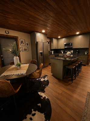 A modern kitchen with wooden cabinets, a refrigerator, a microwave, and a dining area with a table and chairs.