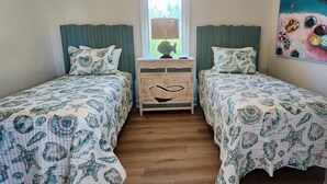 Una habitación con dos camas, cada una con ropa de cama y almohadas a juego, una mesita de noche de madera con cajones, un ventanal con vistas y un cuadro colorido en la pared.