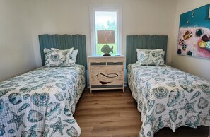 A room with two beds, each with matching bedding and pillows, a wooden nightstand with drawers, a window with a view, and a colorful painting on the wall.