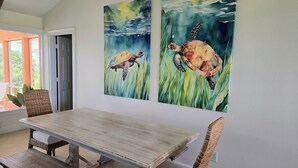 Un comedor con mesa de madera y sillas de mimbre, un ventanal grande con marcos naranja y dos cuadros colgados en la pared de tortugas marinas.