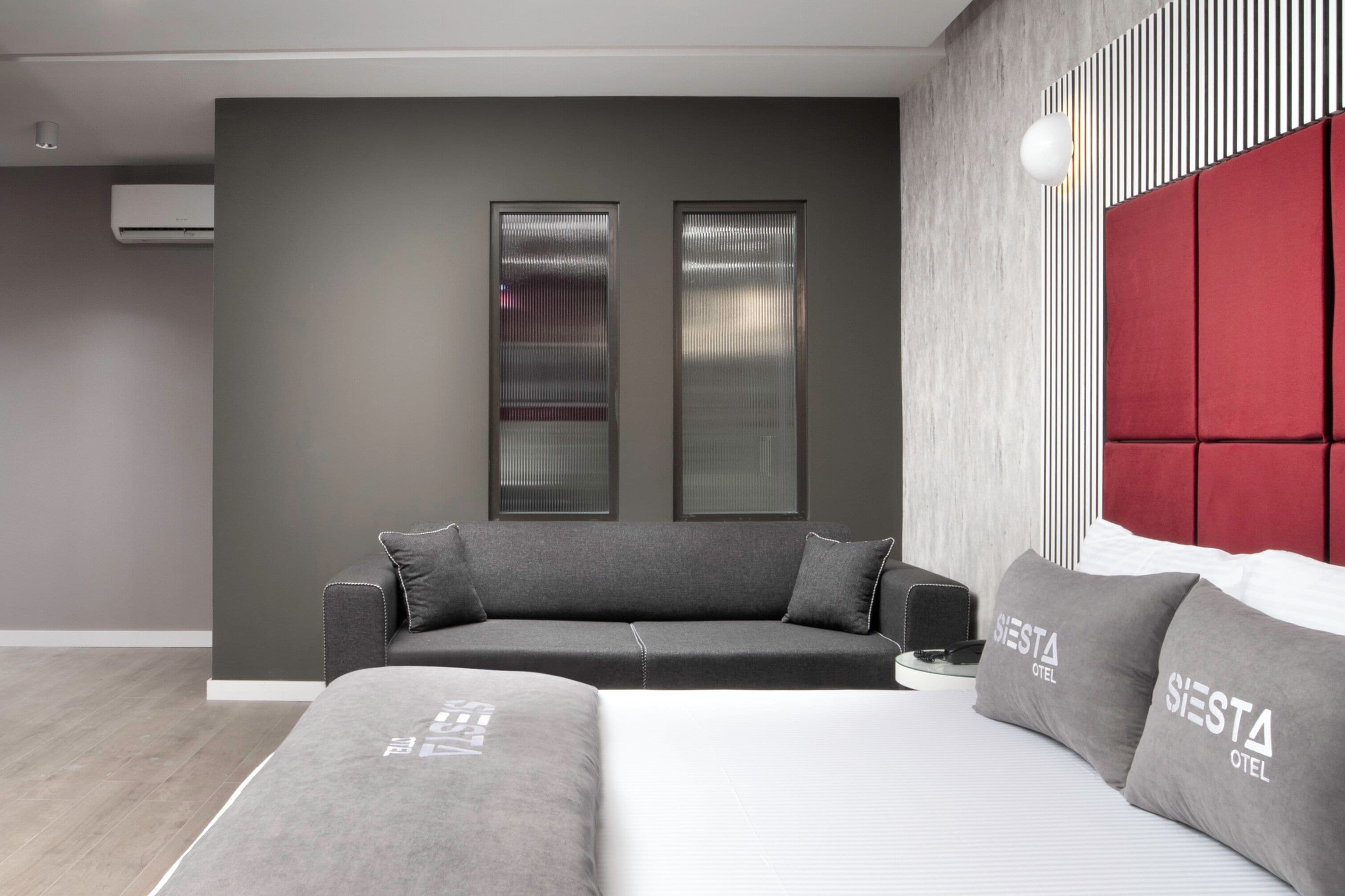 A modern hotel room with a grey sofa, a bed with grey bedding, and a red headboard.