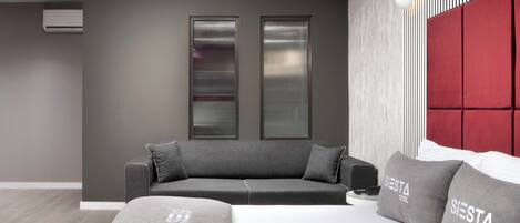 A modern hotel room with a grey sofa, a bed with grey bedding, and a red headboard.