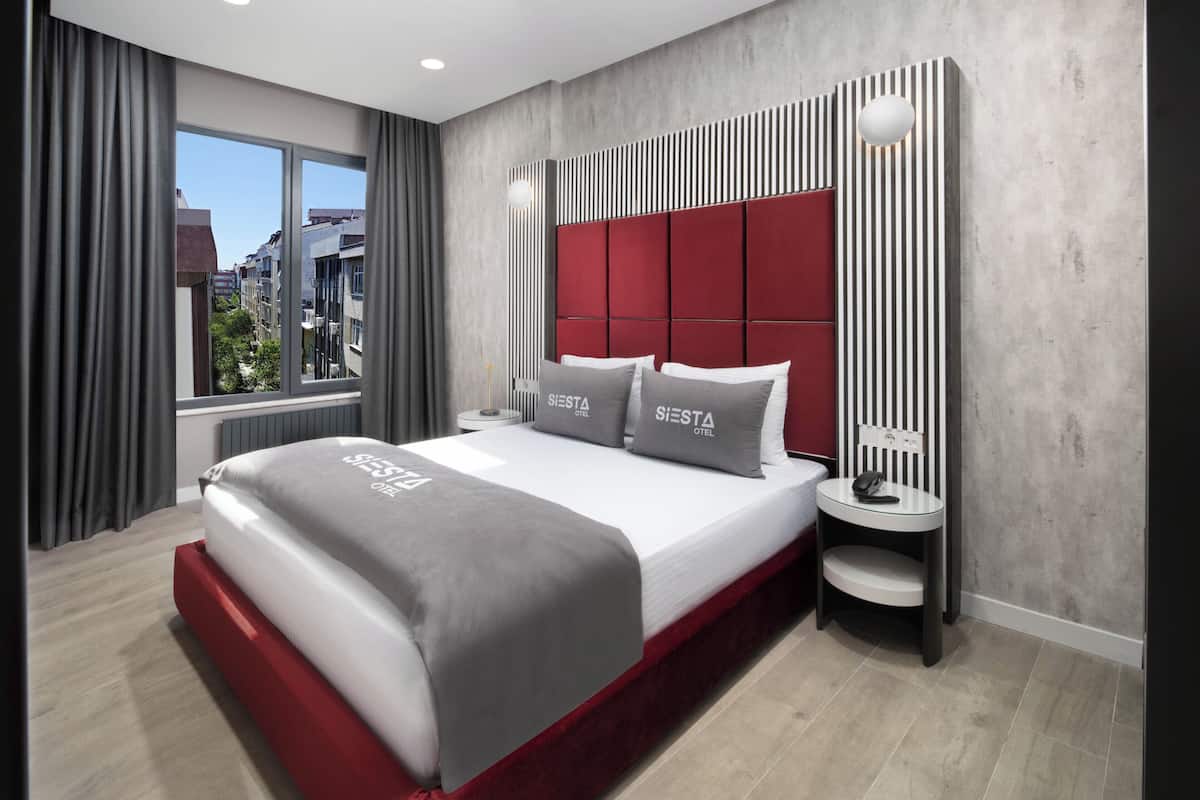 Una habitación de hotel moderna con una cama grande, una mesita de noche, cabecero rojo y una ventana con cortinas.
