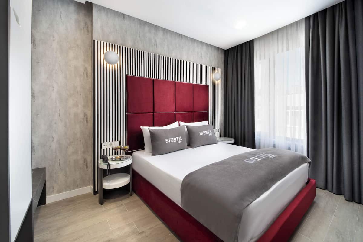 Una habitación de hotel moderna con una cama grande, una mesita de noche con lámpara y vistas a la ciudad a través de cortinas translúcidas.