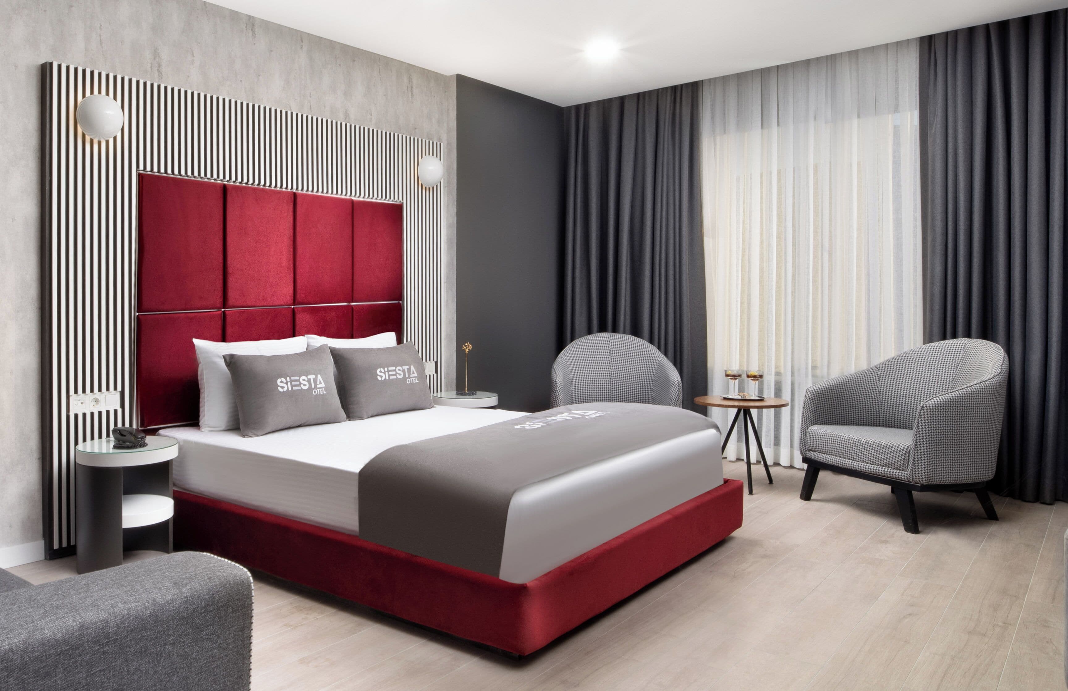 A modern hotel room with a red upholstered headboard, a grey and white bed, a grey armchair, a small round table with a lamp, and a large window with sheer curtains.