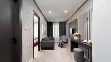 A modern living room with a grey sofa, a round coffee table, and a grey ottoman. There is a flat-screen TV mounted on the wall, a side table with a vase of flowers, and a large window with curtains.