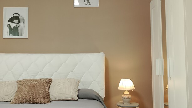 A bedroom with a bed, a nightstand with a lamp, and two framed pictures on the wall. - Adam Venice 1 (Mira)