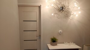 A hallway with a closed door, a small table with a plant and decorative items, and a wall-mounted light fixture. - Adam Venice 1 (Mira)