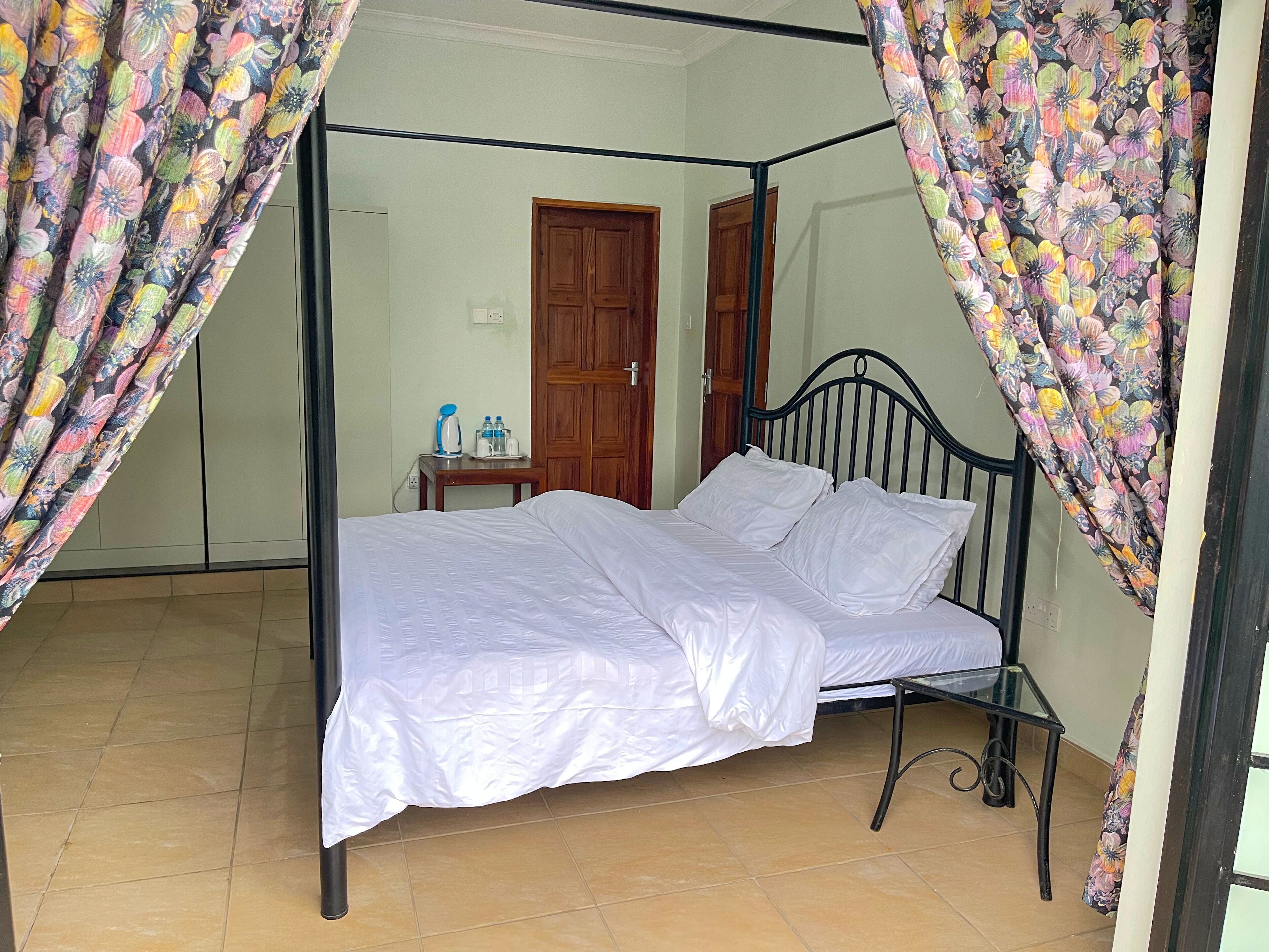 A four-poster bed with white bedding, a wooden nightstand with a kettle and water bottles, and a wooden door in the background.