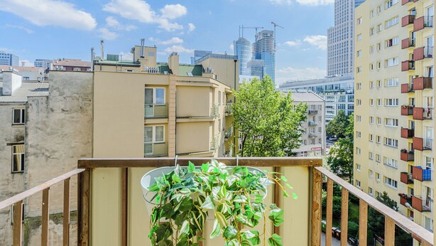A balcony with potted plants, a view of a cityscape with modern buildings, and a clear sky.