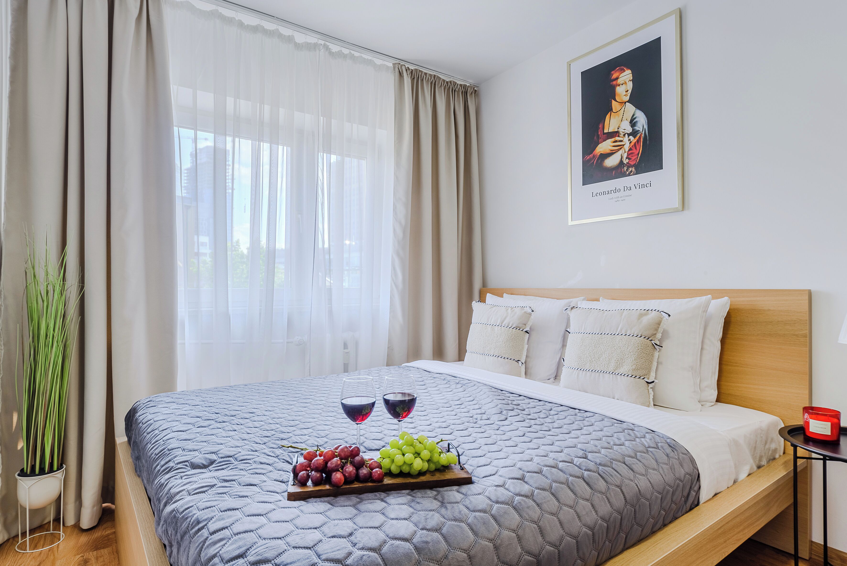 Hotellihuone, jossa on sänky, yöpöytä, jossa on punainen kynttilä, seinällä oleva taideteos ja ikkuna, jossa on läpikuultavat verhot.
