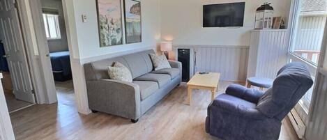A cozy living room with a grey sofa, a wooden coffee table, and a blue armchair. There is a ceiling fan, a flat-screen TV, and artwork on the walls.