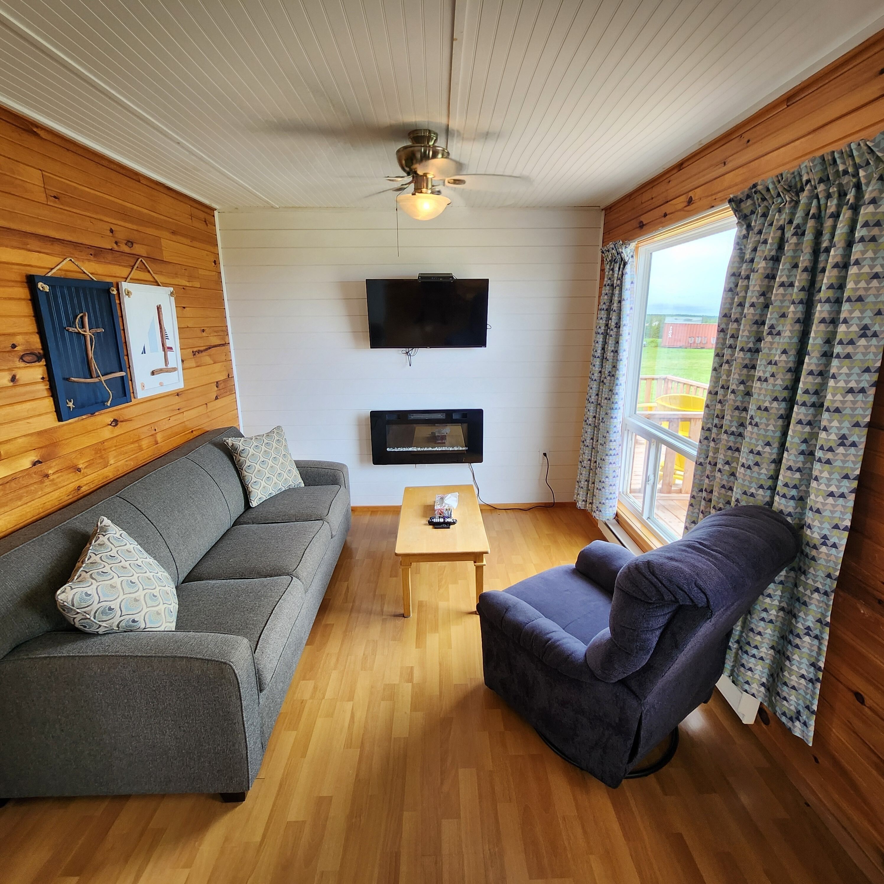 A cozy living room with a grey sofa, a dark armchair, a wooden coffee table, and a flat-screen TV mounted on the wall.