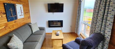 A cozy living room with a grey sofa, a dark armchair, a wooden coffee table, and a flat-screen TV mounted on the wall.