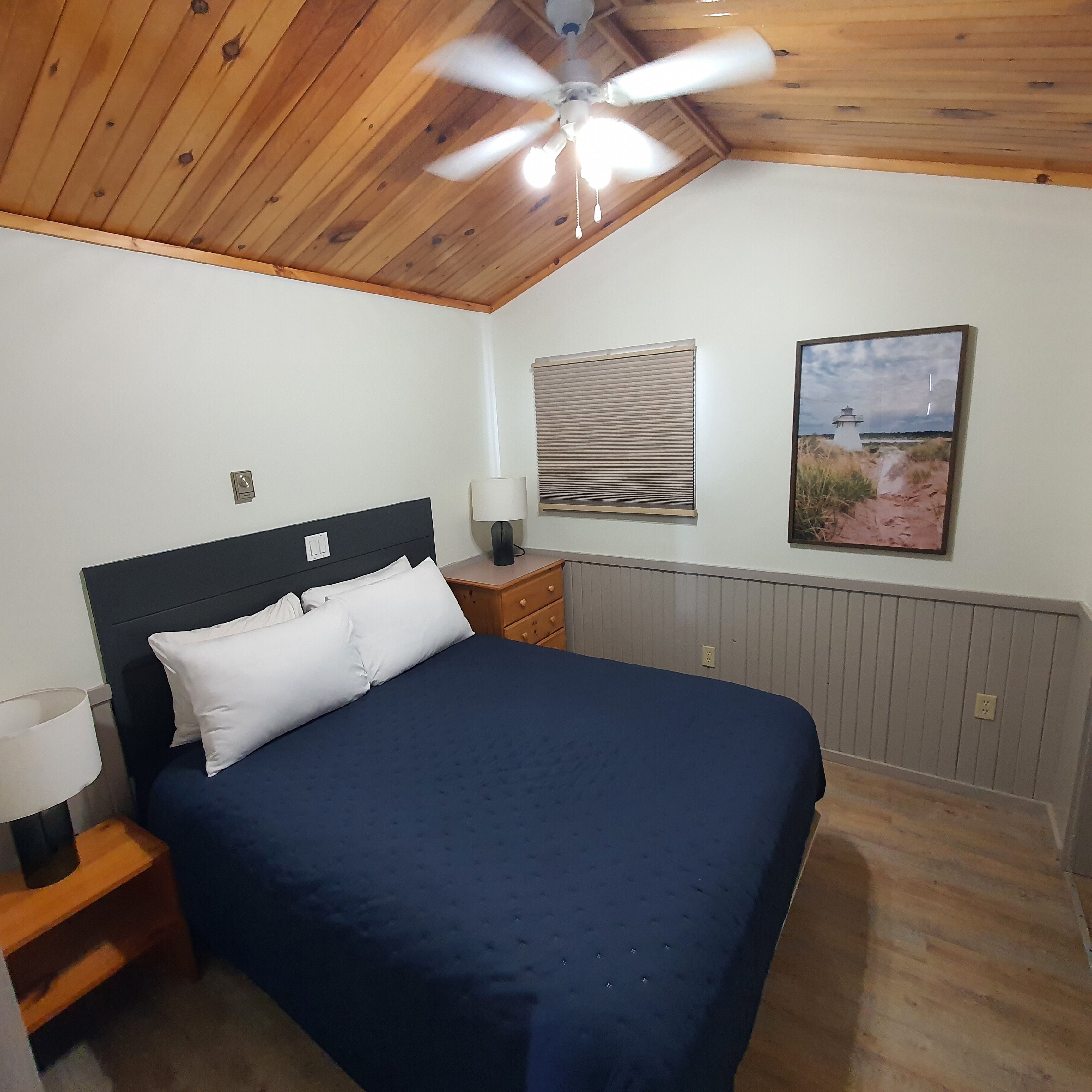 Une chambre à coucher comprenant un lit, une table de chevet, une lampe, un ventilateur de plafond, une fenêtre avec des stores et un tableau encadré accroché au mur.