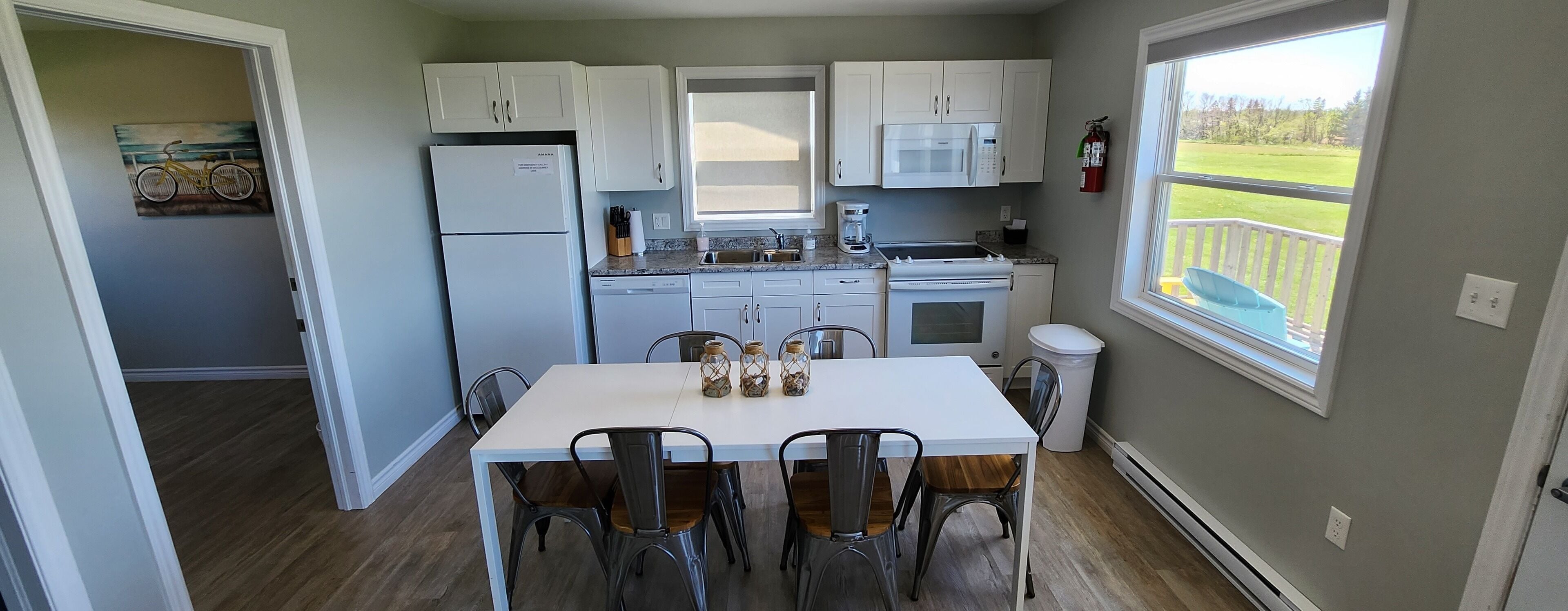 A compact kitchen with white cabinets, a refrigerator, a microwave, and a coffee maker. There is a dining table with chairs and a window offering a view of a grassy area.
