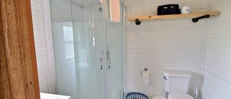 Un baño con ducha de mampara de vidrio, inodoro, lavamanos y un estante con artículos de aseo.