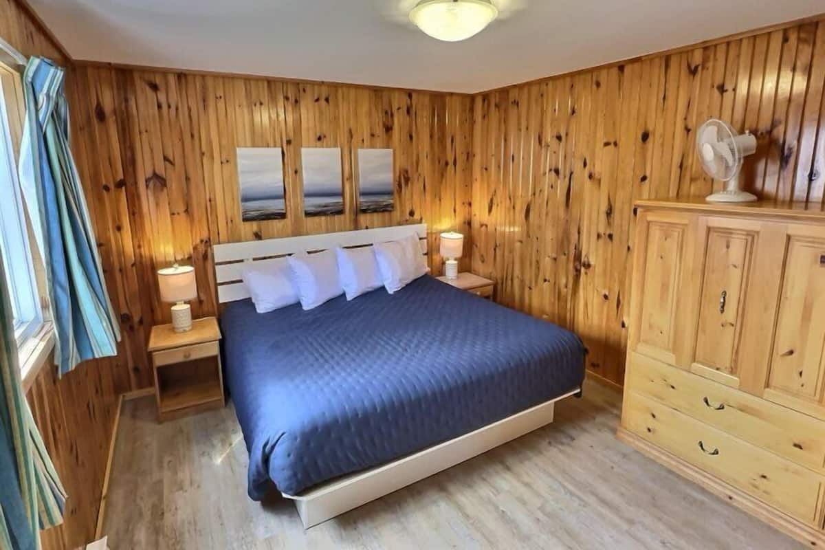 Een slaapkamer met een houten lambrisering, een bed met wit beddengoed, twee nachtkastjes met lampen en een houten dressoir.