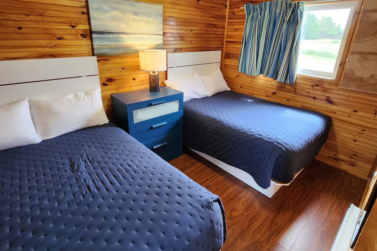 Een kamer in chaletstijl met twee bedden, houten wanden, een schilderij, een nachtkastje, een lamp en een raam met gordijnen.