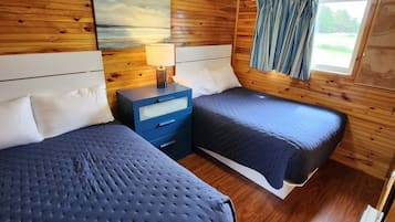 A cabin-style room with two beds, wooden walls, a painting, a nightstand, a lamp, and a window with curtains.