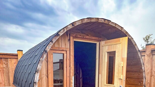Una estructura en forma de barril de madera con una puerta y ventanas, un banco en el exterior y un camino de piedra.