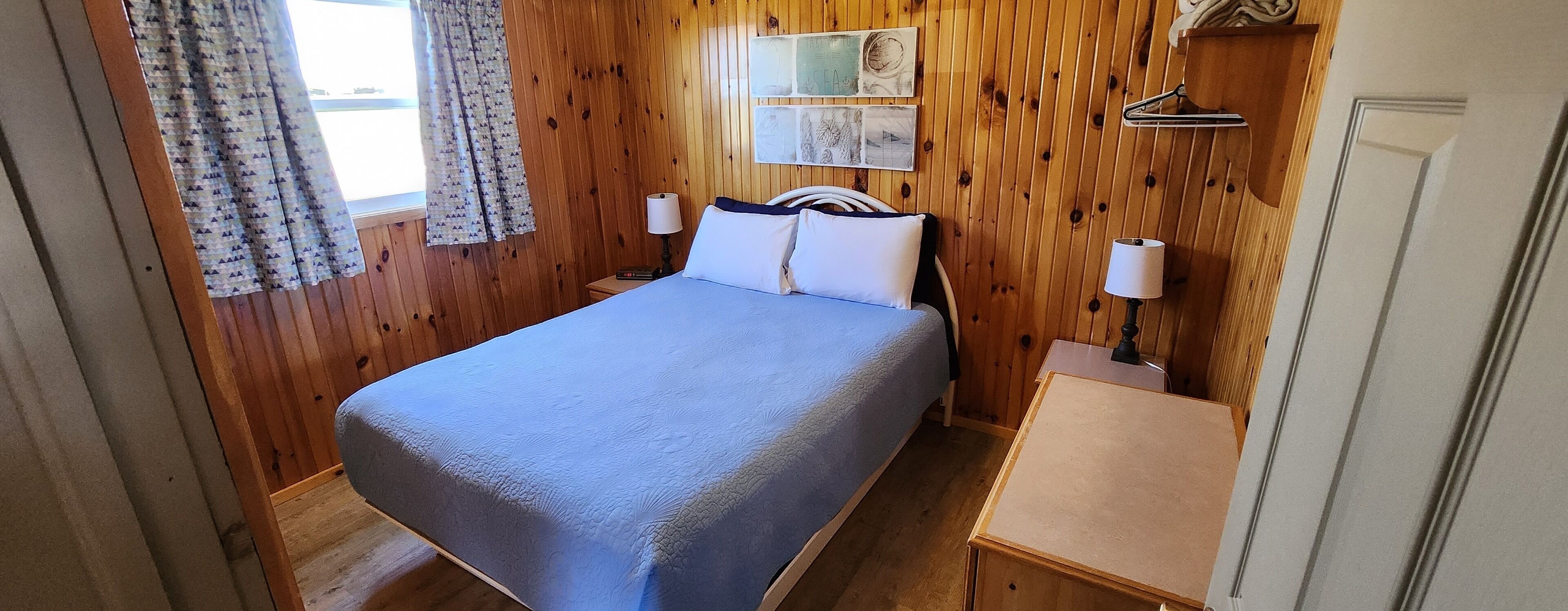 Habitación con paneles de madera, una cama, una mesita de noche pequeña y una estantería en la pared.