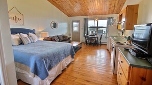 Un dormitorio con una cama grande, una zona de comedor con mesa y sillas, una cocina con estufa y televisión, y un techo de madera.
