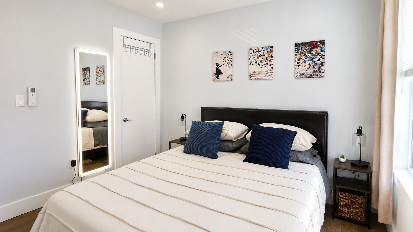 A bedroom with a bed, a nightstand, a lamp, a wall-mounted mirror, and artwork.